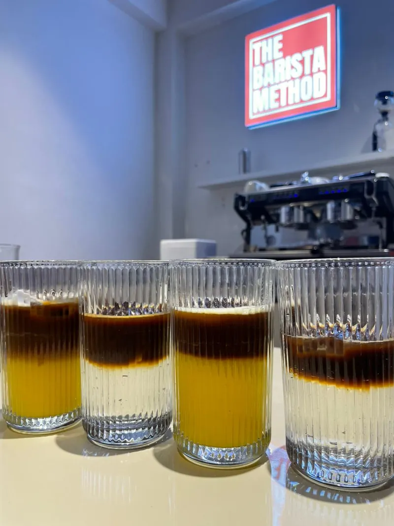 Four layered coffee tonic drinks in ribbed glasses on the lab counter, with The Barista Method sign behind them